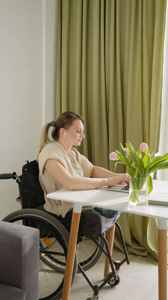 A Woman in Beige Tank Top Sitting on Wheelchair while Using Her Laptop